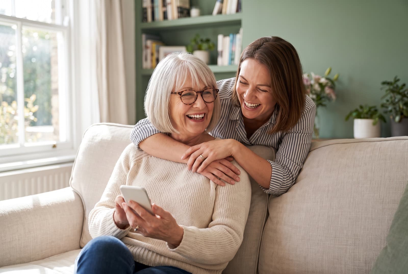 A grandmother laughing on the sofa while her daughter hugs her from behind